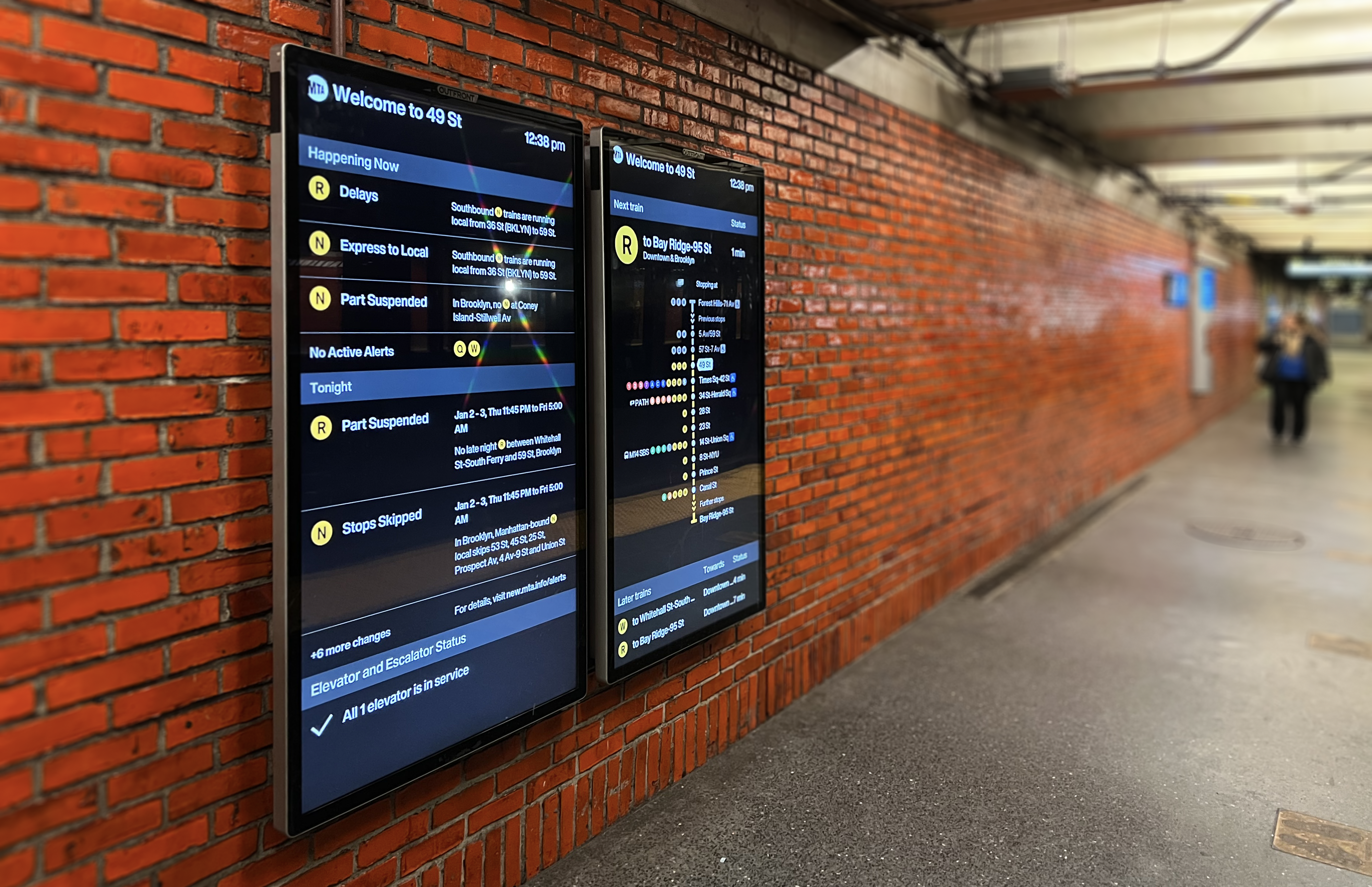 A photo of orange brick wall of the 49th St subway station. There are two vertical screens hanging on the wall, one showing upcoming arrivals and one showing alerts.