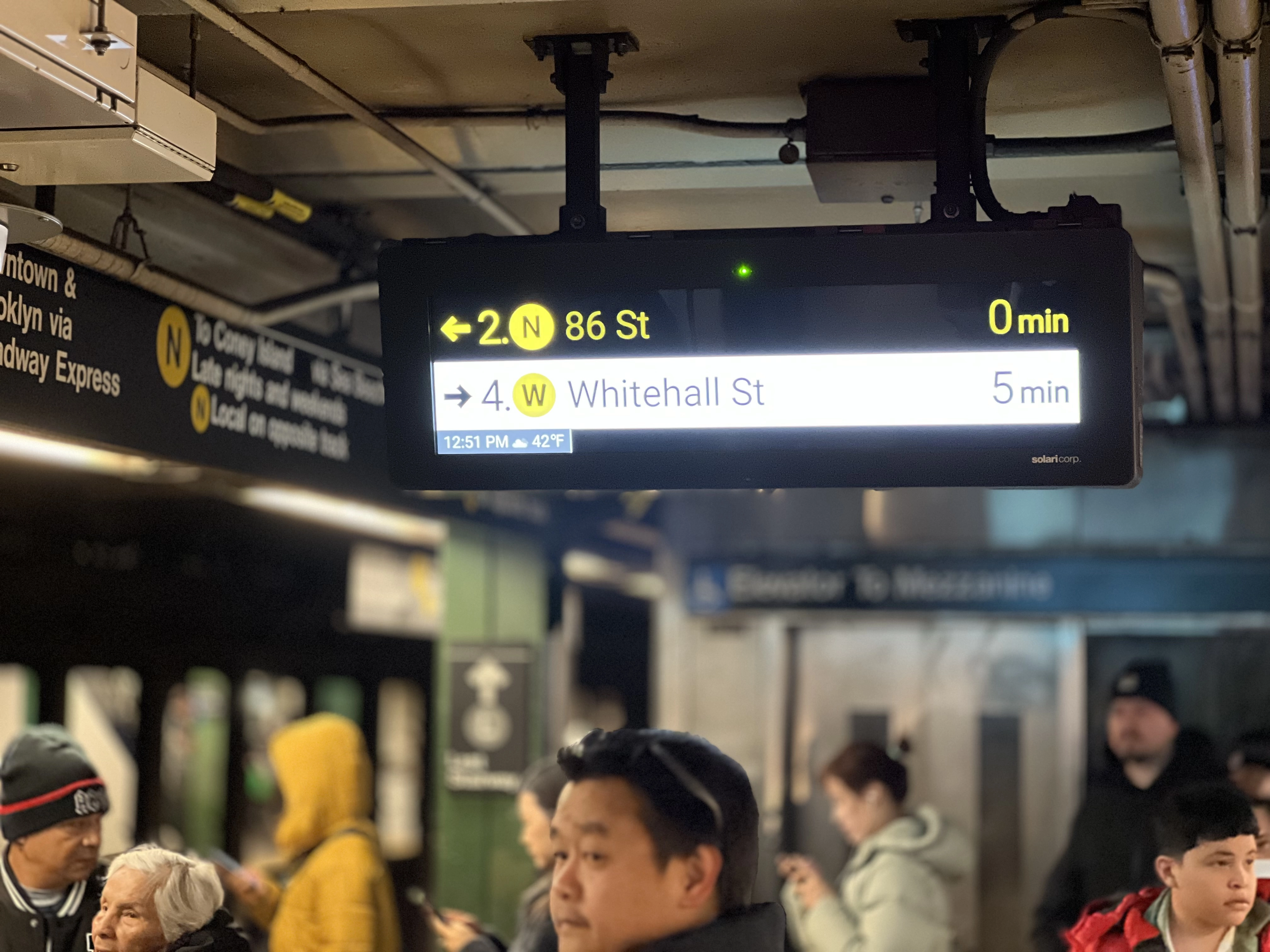 A rectangular screen hanging from the ceiling of a Subway station showing train arrivals.