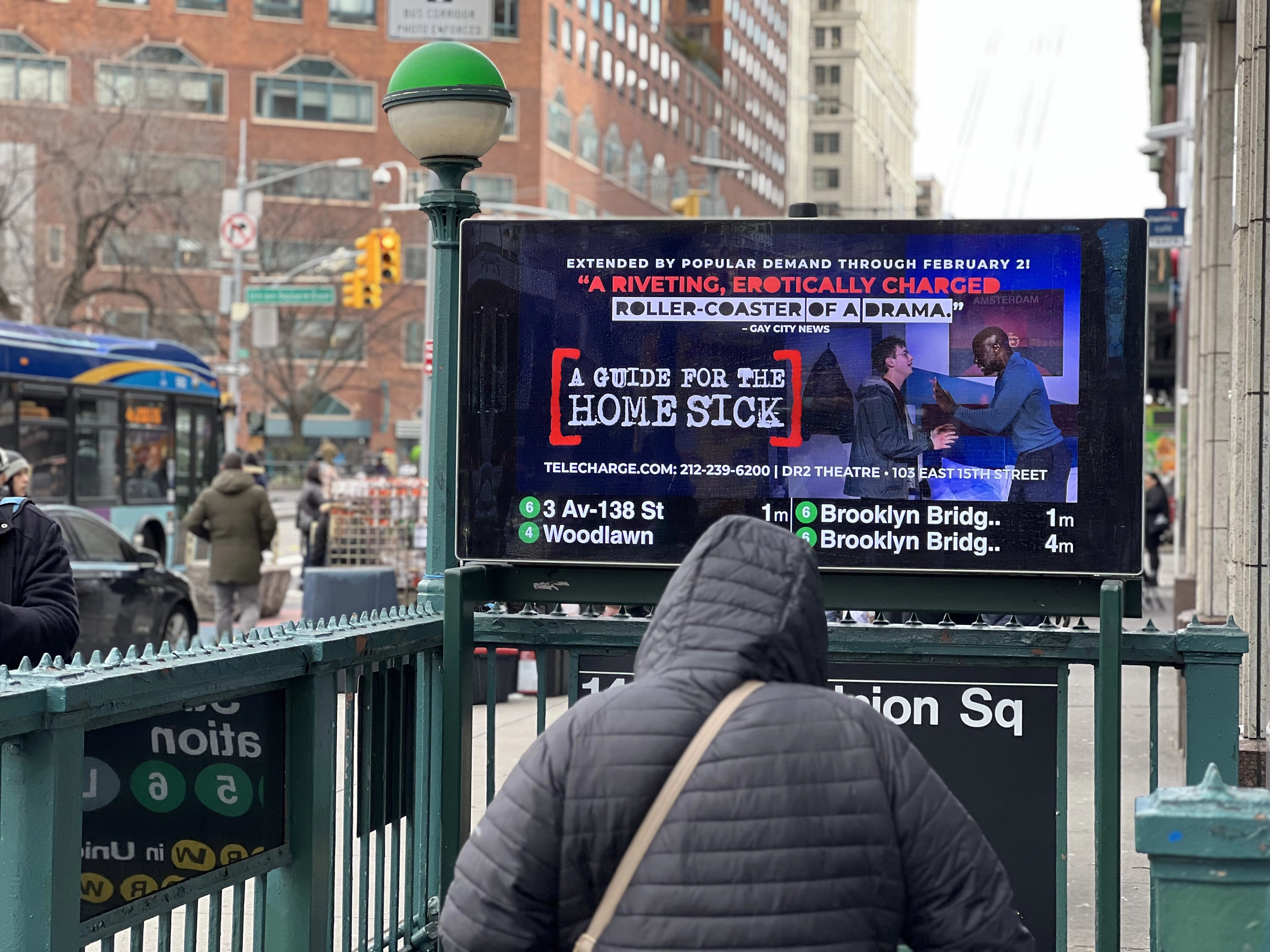 The street-level entrance to a Subway station. A large screen mostly displays an ad, but the bottom few inches show train arrival times.